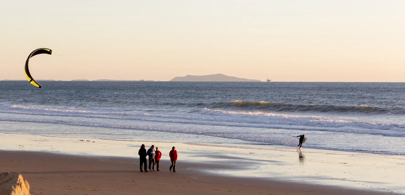 A beach at dusk with people at the waterline and a kite surfer offshore